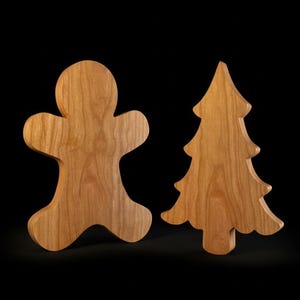 May include: Two wooden Christmas decorations: a gingerbread man and a Christmas tree. Both are cut from light-colored wood with visible grain patterns. The gingerbread man has a classic shape, and the tree has a traditional silhouette.