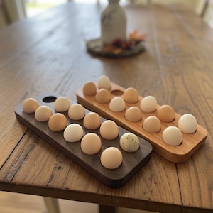 May include: Two wooden egg holders, one dark brown and one light brown, each holding a dozen eggs. The eggs vary in color from light tan to white, with one speckled egg. The holders are on a wooden table.