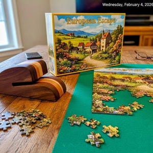 May include: A wooden puzzle holder with a striped design sits on a wooden table next to a partially assembled jigsaw puzzle. The puzzle box, titled "European Escape," depicts a European village scene. A green felt mat is under the puzzle.