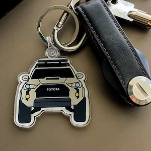 May include: A clear plastic keychain with a cartoon illustration of a white Toyota SUV with a roof rack. The keychain is attached to a black leather key holder with a silver metal loop.