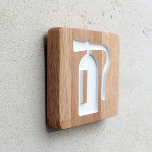 May include: A wooden sign with a white fire extinguisher symbol carved into it. The sign is mounted on a wall.