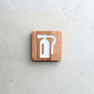 May include: A wooden sign with a white fire extinguisher symbol. The sign is made of light brown wood and has a slightly rough texture.