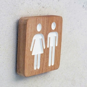 May include: A wooden sign with white silhouettes of a person and a person with a skirt. The sign is a light brown color.