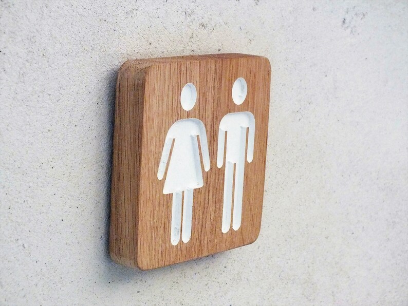 May include: A wooden sign with white silhouettes of a person and a person with a skirt. The sign is a light brown color.