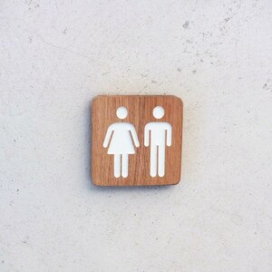 May include: A wooden sign with white painted symbols for a restroom. The sign has a light brown wood grain.