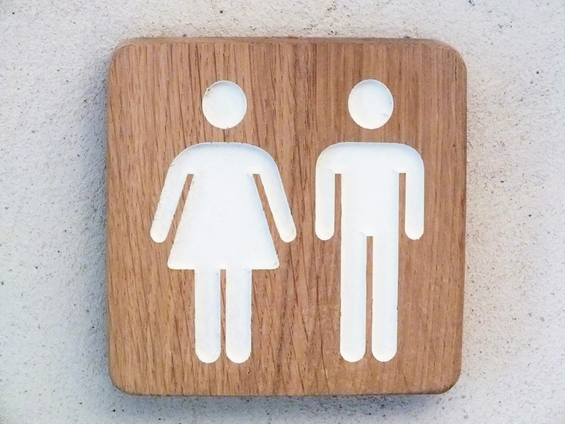 May include: A wooden sign with white painted symbols of a person and a woman. The sign is a light brown color with a wood grain pattern.