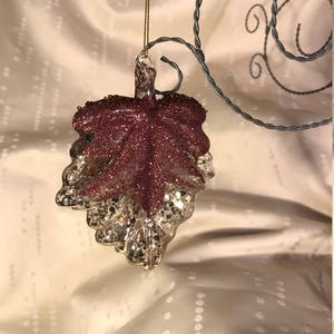 May include: A glass leaf ornament with a glittery, metallic finish. The leaf is a deep burgundy color with a silver glitter overlay.