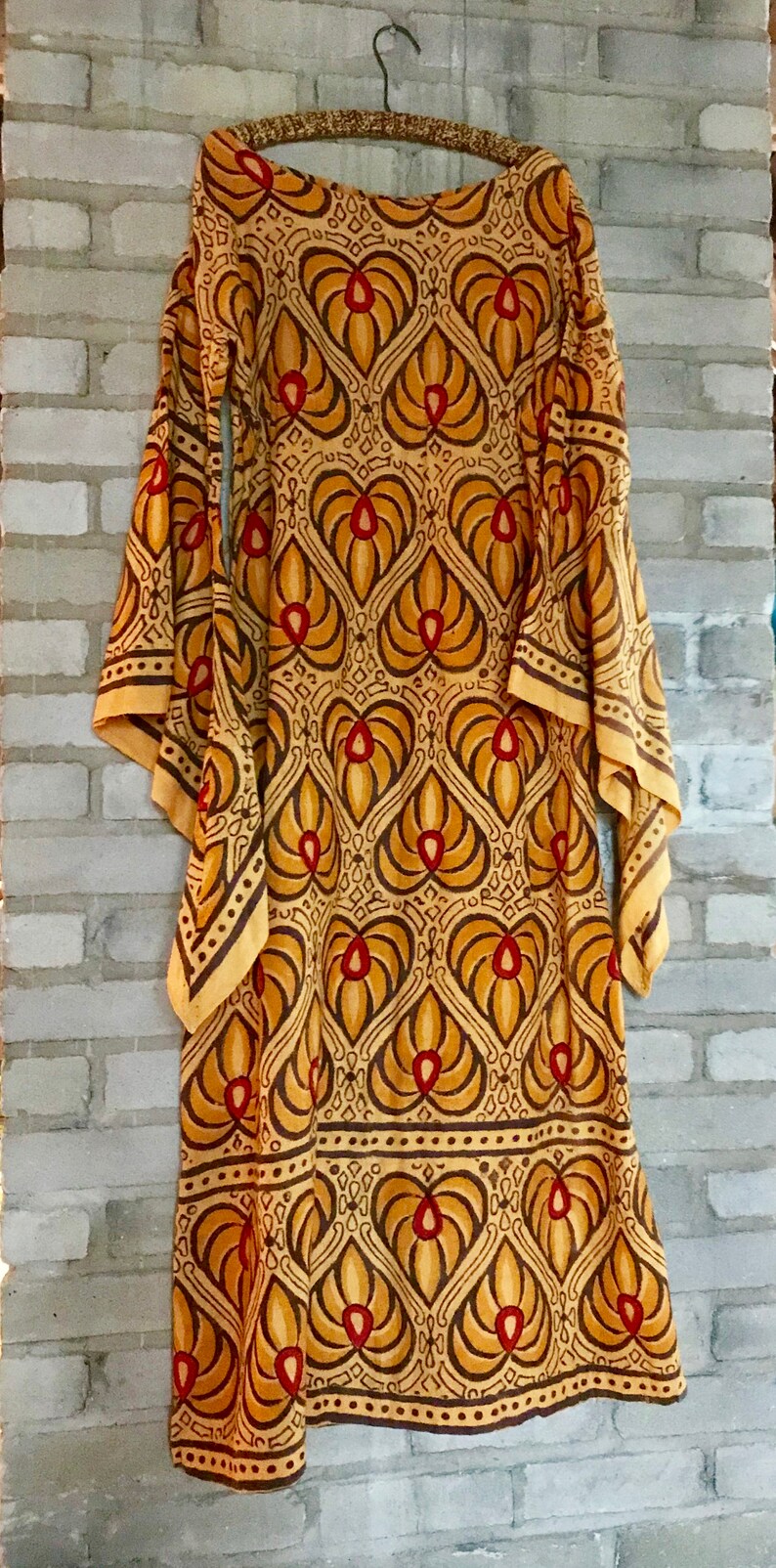 May include: A long, yellow dress with a brown and red geometric pattern. The dress has a scoop neck and long sleeves. The sleeves are wide and bell-shaped.