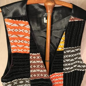 May include: A black sleeveless vest with a patchwork design. The vest is made of different fabrics with geometric patterns in orange, brown, white, and black. The vest has a satin lining and a label that reads "Zora Handmade India".