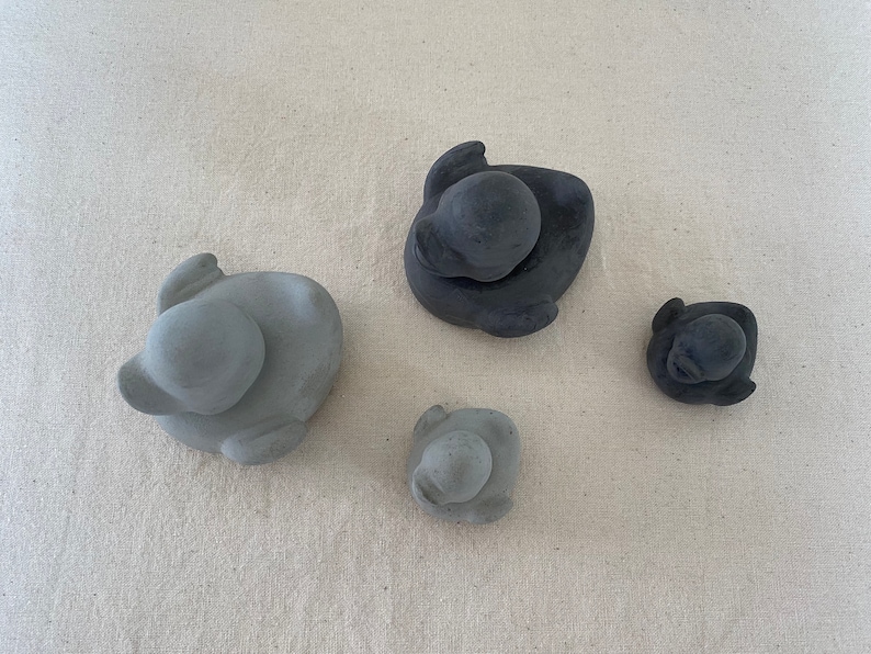 Może przedstawiać: Cztery małe ceramiczne figurki kaczek. Dwie są szare, a dwie czarne. Kaczki siedzą wszystkie w pozycji siedzącej z pochyloną głową.