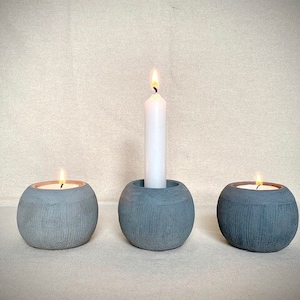 Könnte beinhalten: Drei graue Beton-Kerzenhalter mit einer weißen Kerze in Form einer Säule im mittleren Halter.