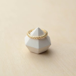 Puede incluir: Un anillo de oro con un diseño tejido se encuentra sobre un soporte de exhibición geométrico blanco.