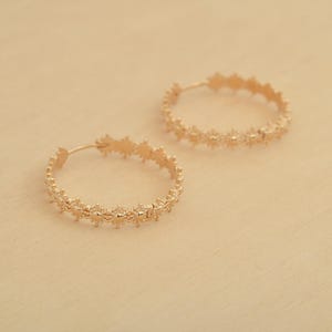 以下が含まれることがあります： 繊細で複雑なデザインのゴールドのフープピアスのペア。フープは薄い金属製で、小さな星形のディテールが繰り返されるパターンが特徴です。