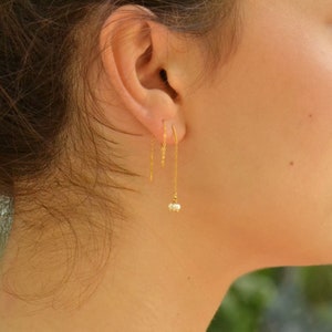 Może przedstawiać: Złote kolczyki z łańcuszkiem i małym gronem pereł na końcu. Kolczyki wiszą z płatka ucha.