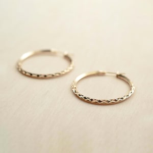 May include: Two gold-colored hoop earrings with a textured, diamond-cut pattern. The earrings are displayed on a light beige surface. The earrings are a classic jewelry item.