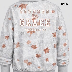 Könnte beinhalten: Ein hellgraues Sweatshirt mit floralem Muster und den Worten "COVERED by GRACE" in braunen Buchstaben. Der Text "Ephesians 2:8" steht unter dem Wort "GRACE". Die Rückseite des Sweatshirts ist sichtbar.