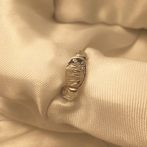 May include: A silver ring with a textured band and an oval-shaped top. The top of the ring has a detailed design.
