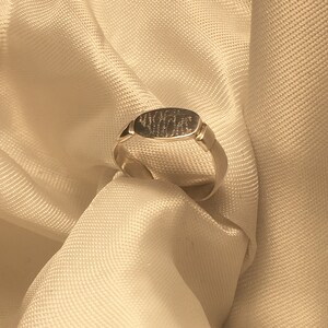 May include: A silver ring with a rectangular band and a flat oval top. The top of the ring has an inscription.