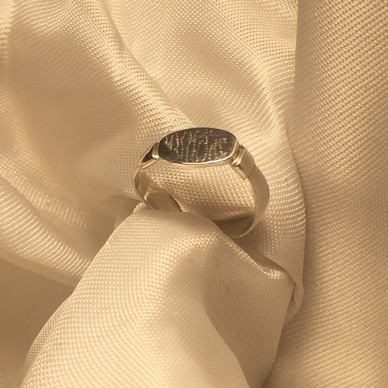 May include: A silver ring with a rectangular band and a flat oval top. The top of the ring has an inscription.