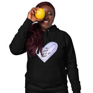May include: Black hoodie featuring a white and light purple heart design with the text "Feed the Algorithm with all your HEART". A person is holding a yellow lemon in front of their face.