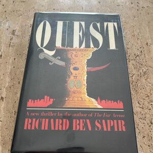 May include: A black hardcover book with the title "QUEST" in large white letters. The cover features a gold chalice with two daggers crossed behind it. The book is by Richard Ben Sapir and is described as "A new thriller by the author of "The Far Arena."