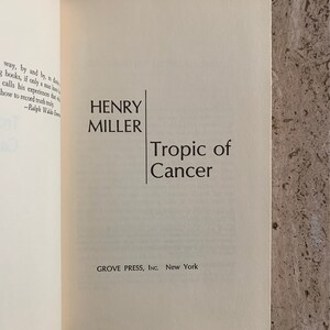 May include: A white book with black text on the cover. The title is "Tropic of Cancer" by Henry Miller. The book is published by Grove Press, Inc. in New York.