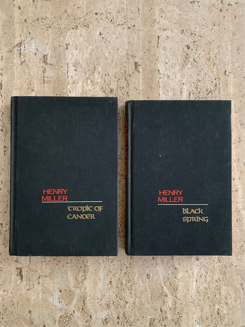 May include: Two black hardcover books with gold lettering. The book on the left is titled "Tropic of Cancer" by Henry Miller. The book on the right is titled "Black Spring" by Henry Miller.