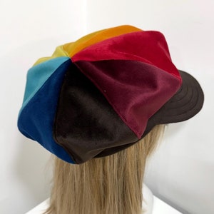 May include: A multi-colored newsboy cap featuring panels of velvet fabric in shades of red, orange, yellow, light blue, dark blue, and dark brown. The cap has a structured dark brown brim with visible stitching and a soft, rounded crown shape.