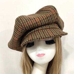 Peut inclure: Casquette de style gavroche en tweed marron et vert avec un motif à chevrons. La casquette a une couronne arrondie et une petite visière rigide. Elle est présentée sur une tête de mannequin avec des cheveux blonds.