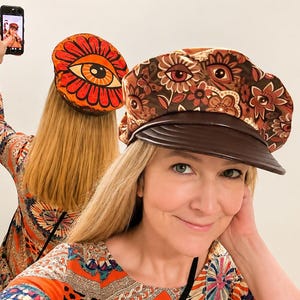 May include: A baker boy cap featuring a brown fabric with a multi-coloured floral and eye pattern, paired with a dark brown brim. The cap is worn by an individual who is also wearing a matching long-sleeved top with a vibrant, intricate print.