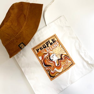 May include: A white canvas tote bag with a brown corduroy bucket hat on top. The tote bag features a graphic design of a woman with long brown hair blowing in the wind. The graphic includes the text "People are strange" and "You'll meet them on the way".