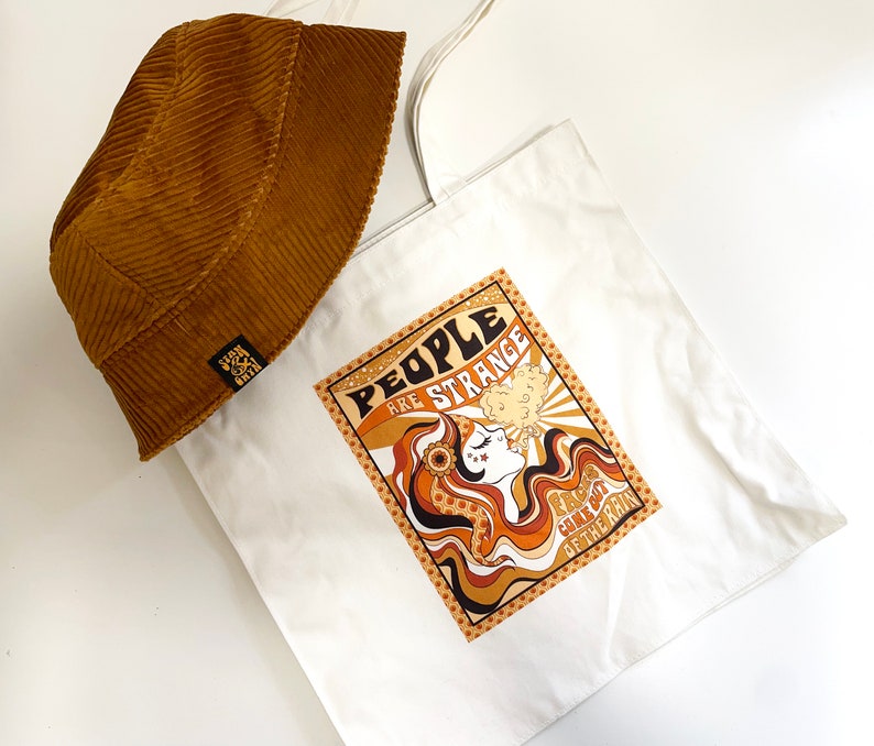 May include: A white canvas tote bag with a brown corduroy bucket hat on top. The tote bag features a graphic design of a woman with long brown hair blowing in the wind. The graphic includes the text "People are strange" and "You'll meet them on the way".