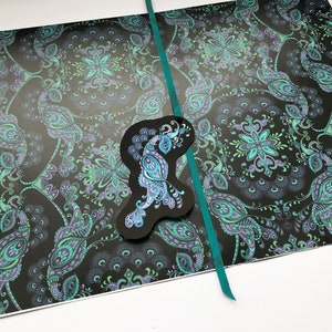 K&ouml;nnte beinhalten: Ein schwarzes Papier mit einem Muster in Teal und einem ausgeschnittenen Pfauenmotiv. Das Papier ist in der Mitte gefaltet und mit einem Teal-Band zusammengebunden.