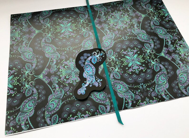 K&ouml;nnte beinhalten: Ein schwarzes Papier mit einem Muster in Teal und einem ausgeschnittenen Pfauenmotiv. Das Papier ist in der Mitte gefaltet und mit einem Teal-Band zusammengebunden.