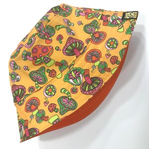 May include: A yellow bucket hat with a mushroom print. The mushrooms are pink, green, and orange with a psychedelic design. The hat has an orange underside.