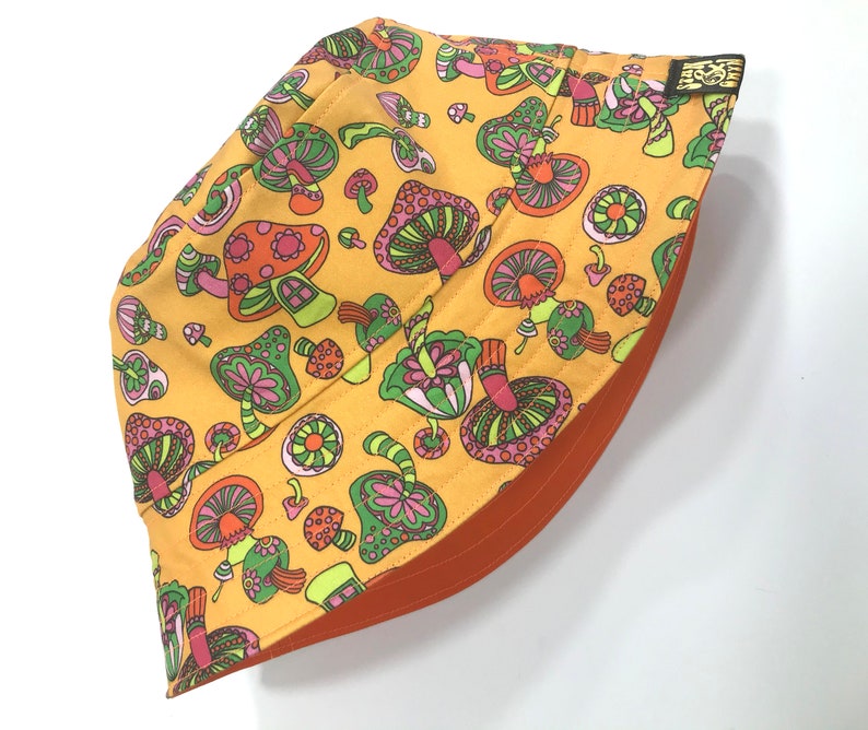 May include: A yellow bucket hat with a mushroom print. The mushrooms are pink, green, and orange with a psychedelic design. The hat has an orange underside.