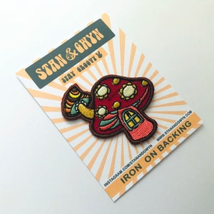 Op de afbeelding: Strijk-op patch met een rode en witte paddenstoel met een klein huisje op de bovenkant. De patch heeft een bruine omtrek en bevindt zich op een witte kaart met de tekst "Stan & Ghywn Stay Groovy" en "Iron on Backing" in zwart.