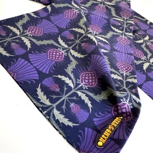 May include: A navy blue silk scarf with a repeating pattern of purple thistles. The scarf has a gold label with the text "OXFORD & JAMES" on it.