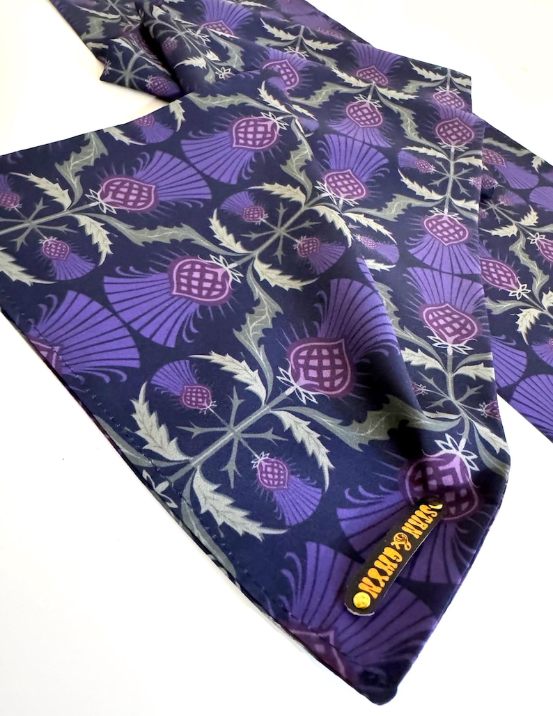 May include: A navy blue silk scarf with a repeating pattern of purple thistles. The scarf has a gold label with the text "OXFORD & JAMES" on it.