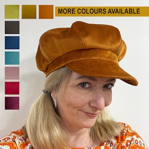 Puede incluir: Una gorra de boina de terciopelo marrón con una visera ancha. La gorra la lleva puesta una persona con el pelo rubio y está peinada con dos coletas. El texto "MÁS COLORES DISPONIBLES" se muestra encima de la gorra.