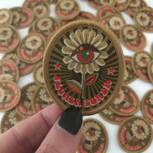 Könnte beinhalten: Nahaufnahme eines gestickten Aufnähers in Braun und Beige mit einer stilisierten Blume mit einem Auge in der Mitte. Die Blume hat eine rote Mitte und grüne und goldene Akzente. Der Text "Flower Power" ist in roter, kursiv geschriebener Schrift um die Blume herum geschrieben.