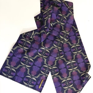 May include: A purple and green patterned silk scarf with a thistle design. The scarf has a gold label that reads "Charlie & Max".