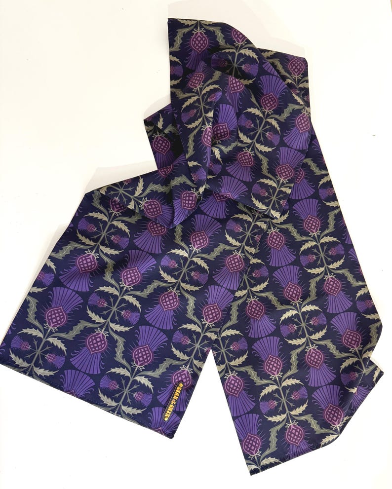 May include: A purple and green patterned silk scarf with a thistle design. The scarf has a gold label that reads "Charlie & Max".