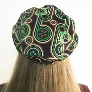 Puede incluir: Un gorro negro con un patrón geométrico verde, marrón y blanco. El patrón presenta círculos, espirales y flores.