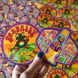 May include: Two colourful stickers with a groovy 70s theme. The first sticker features a green frog sitting on a lily pad with the text "Totally Groovy" in a rainbow font. The second sticker features a lava lamp with the text "Gotta Lava Whole Love" in a rainbow font.