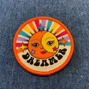 May include: A round orange patch with a white background and a colourful sun with a face and the word "Dreamer" in black letters.