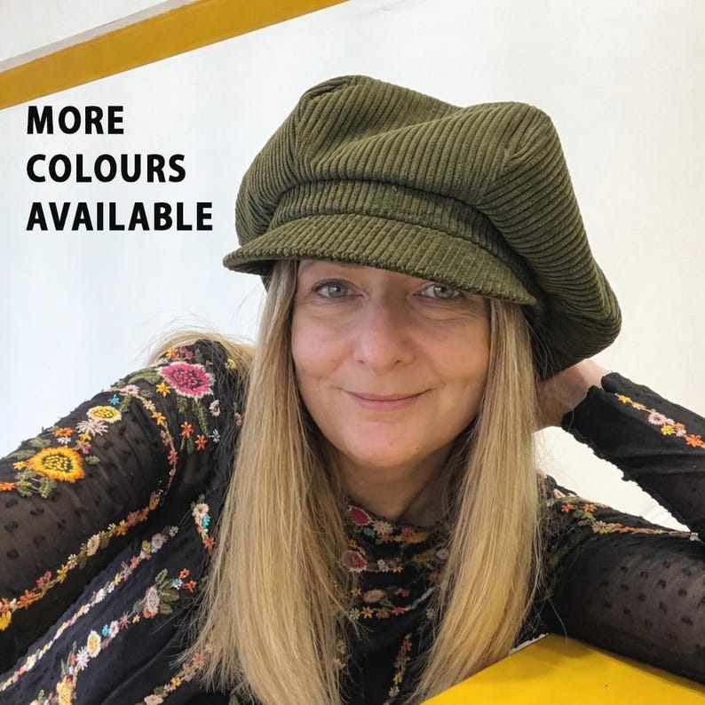 May include: An olive green corduroy newsboy cap with a structured brim and a soft, rounded crown. The text "MORE COLOURS AVAILABLE" is printed in black on the upper left side of the image, indicating additional options for this headwear item.