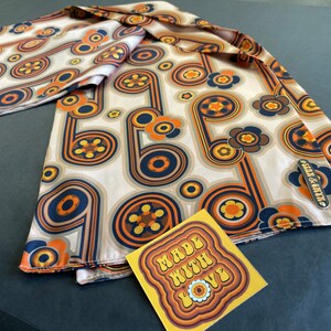 Pu&ograve; includere: Un foulard di seta beige con un motivo retr&ograve; di cerchi e fiori arancioni, gialli e blu. Il foulard ha un'etichetta che dice "Silk & Satin".