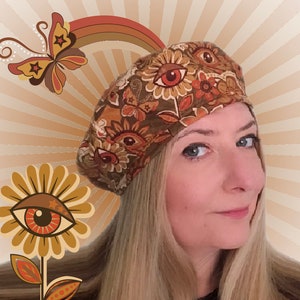 May include: A brown beret with a floral and eye pattern is worn by a person with blonde hair. The beret has a retro design with orange, brown, and white colors. The background features a butterfly, rainbow, and flower with an eye design.