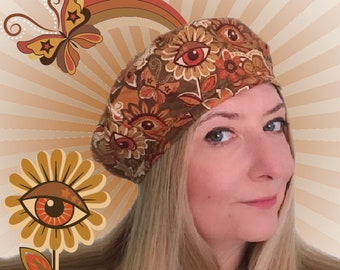 May include: A brown beret with a floral and eye pattern is worn by a person with blonde hair. The beret has a retro design with orange, brown, and white colors. The background features a butterfly, rainbow, and flower with an eye design.